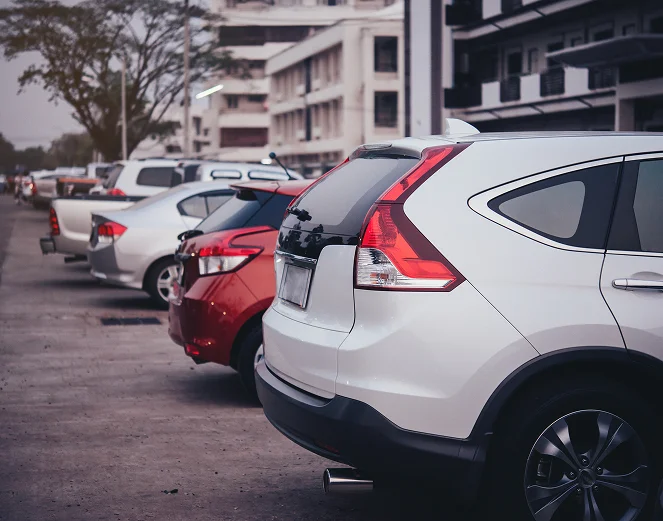 A imagem mostra um parque de estacionamento na rua com vários automóveis estacionados, de diferentes modelos e cores, alinhados ao longo do passeio.
