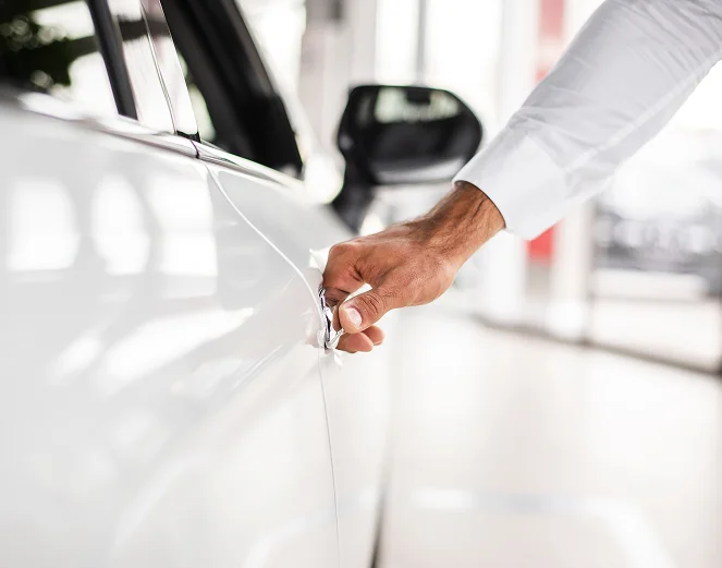 Na imagem, vê-se um homem de pé no interior de um concessionário automóvel moderno ou de um parque de estacionamento espaçoso.