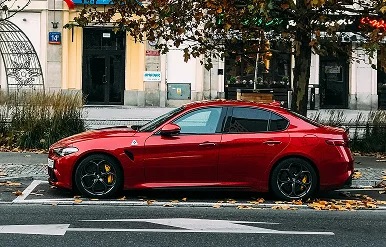 No centro da imagem encontra-se um elegante sedan desportivo vermelho, com vidros escurecidos e uma distância ao solo reduzida. O carro está estacionado paralelamente ao passeio; na parte dianteira, são visíveis jantes de liga leve pretas com detalhes em amarelo vivo e pneus de perfil baixo.